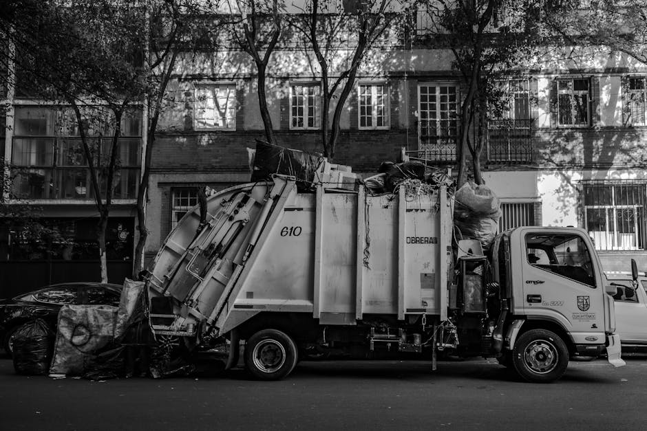 A city street scene featuring a mix of historic and modern buildings. In the foreground, a small waste collection truck with a white and blue cabin is parked on the side of the road, appearing to be i