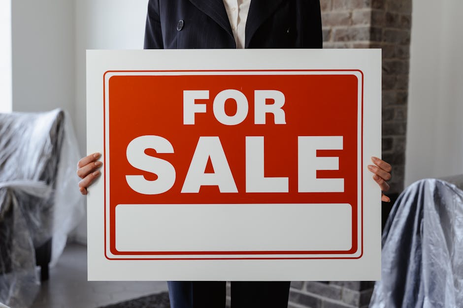 A person wearing a dark jacket is holding a large rectangular sign with a white background and a red border, displaying the words 'FOR SALE' in bold white and red capital letters. The person's face is